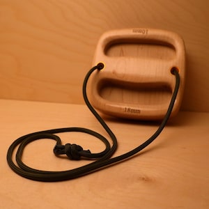 May include: A light-colored wooden climbing hangboard with two horizontal cutouts. The top edge is engraved with 10mm and the bottom edge with 18mm. A black cord is threaded through two holes in the wood and tied in a knot at the bottom.
