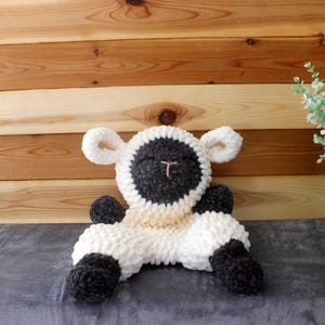 May include: A handmade, plush sheep toy in white, black, and tan. The sheep has a black face and feet, white body, and tan accents. The toy is sitting on a gray surface in front of a wooden background.
