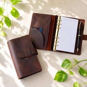 May include: A brown leather planner with a buckle closure, open to reveal a notepad and gold-coloured binder rings. A black pen rests in a holder. A credit card is visible in a pocket. The planner is on a light-coloured surface with green leaves.
