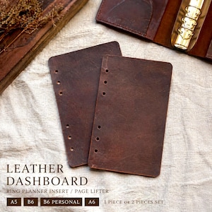 Könnte beinhalten: Zwei braune Leder-Dashboard-Einsätze für Ringplaner. Die Einsätze haben mehrere Löcher zum Binden und sind rechteckig mit abgerundeten Ecken. Der Text auf dem Bild lautet "LEATHER DASHBOARD" und "RING PLANNER INSERT / PAGE LIFTER".