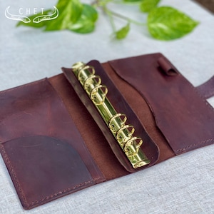 Può includere: Raccoglitore in pelle bordeaux aperto, con anelli color oro. Il raccoglitore è aperto, rivelando una tasca sul lato sinistro e un portapenne. Il logo Chet è visibile nell'angolo in alto a sinistra. Lo sfondo include foglie verdi.