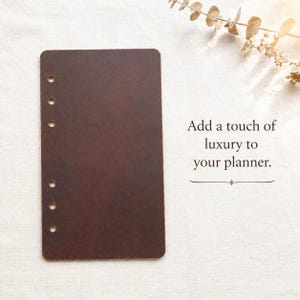 May include: A rectangular dark brown leather planner insert featuring six circular punched holes along the left edge. The text "Add a touch of luxury to your planner." is printed to the right of the leather sheet in a dark serif font.
