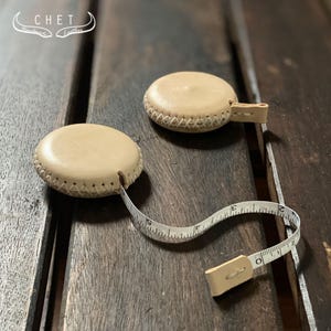 以下が含まれることがあります： ベージュ色の革製メジャー2つ。白いメジャーテープが付いています。メジャーは丸く、縁はステッチで、小さな革製のタブが付いています。1つのメジャーはテープが伸びており、センチメートル単位の測定値が表示されています。「CHET Handmade Leather」のロゴが左上にあります。