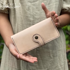 May include: A pale beige leather wallet with black stitching and a button clasp. The rectangular wallet is held by hands. The word "CHET" is visible in the upper left corner of the image.