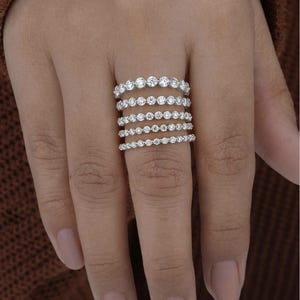 May include: A hand displays six silver rings, each adorned with a series of small, round, clear gemstones. The rings vary in size, creating a layered effect. The gemstones sparkle, catching the light. The background is a warm, neutral tone.