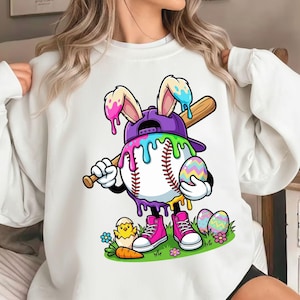 May include: White sweatshirt featuring a baseball character with bunny ears, a purple cap, and a baseball bat. The character holds a colorful Easter egg and is wearing pink sneakers. Easter-themed design with eggs, a chick, and a carrot.