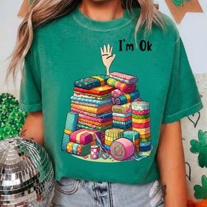 May include: Green t-shirt with a colorful illustration of stacked fabric rolls, bundles, and sewing supplies. The text "I'm Ok" is above the fabric. The design features various patterns and colors, including floral and geometric designs.
