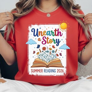 Op de afbeelding: Rood t-shirt met een grafisch ontwerp met de woorden "Unearth a Story" in kleurrijke letters. Het ontwerp bevat een open boek met bloemen, vlinders, wolken en een zon. De tekst "Summer Reading 2026" is ook opgenomen.