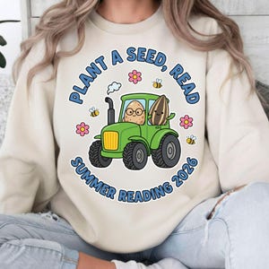 Op de afbeelding: Crèmekleurige sweatshirt met een groene tractorafbeelding. De tractor heeft een aardappel met een bril en een zonnebloem als passagier. Tekst luidt "PLANT A SEED, READ SUMMER READING 2026" met bijen en bloemen.
