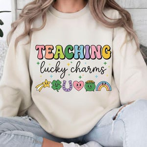 May include: Cream-colored sweatshirt with the text "TEACHING lucky charms" in colorful, retro-style lettering. Below the text are cartoon graphics of a shooting star, shamrock, horseshoe, heart, pot of gold, and rainbow.