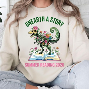 Könnte beinhalten: Beiges Sweatshirt mit einem Grafikdesign eines mit Blumen geschmückten Dinosaurierskeletts, das auf einem offenen Buch steht. Der Text "UNEARTH A STORY" steht über dem Dinosaurier, und "SUMMER READING 2026" darunter.