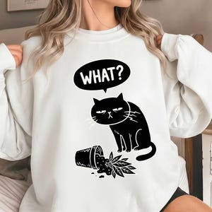 Könnte beinhalten: Weißes Sweatshirt mit einer schwarzen Grafik einer mürrischen Katze und dem Wort "WHAT?" in einer Sprechblase. Die Katze steht neben einem umgestürzten Blumentopf mit verschütteten Blättern. Ein humorvolles Design.