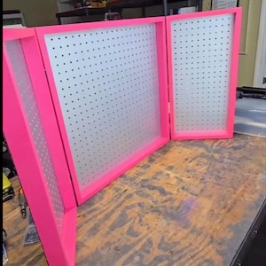 May include: A bright pink, three-panel display board with a pegboard design. The frames are a vibrant pink, and the pegboard surface is a light gray. The display is open, showing the interior pegboard surface. The display is on a wooden surface.