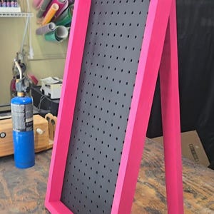 Pode incluir: Um expositor em forma de A rosa brilhante com um centro de placa perfurada cinza escuro. A moldura é feita de madeira e tem um acabamento liso. A placa foi projetada para ficar em pé e tem um suporte de metal preto na parte de trás.