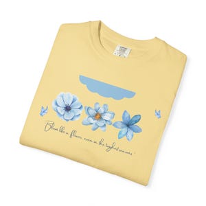 Könnte beinhalten: Hellgelbes T-Shirt mit floralem Design. Es zeigt drei blaue Blumen, eine blaue Wolke und zwei Schmetterlinge. Der Text lautet "Bloom like a flower, even in the toughest seasons". Das Design ist auf die Vorderseite des T-Shirts gedruckt.