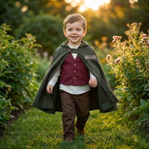 Könnte beinhalten: Ein Kind in einem grünen Kapuzenmantel, einer weinroten Weste, einem weißen Hemd und einer braunen Hose. Der Umhang hat einen Knopf und ein Namensschild mit der Aufschrift "Asher". Das Kind lächelt, während es auf einem grasbewachsenen Pfad geht.