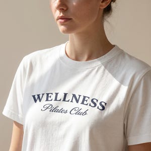May include: White crew neck t-shirt with the words "WELLNESS Pilates Club" printed in navy blue. The shirt has short sleeves and a classic fit, suitable for casual wear or workout sessions.