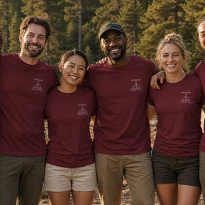 Peut inclure: Un groupe de six personnes portant des t-shirts à manches courtes bordeaux assortis. Chaque t-shirt présente un petit motif blanc sur la poitrine gauche représentant une tente et le texte "TRAILBLAZER CAMP CLUB" en police simple.
