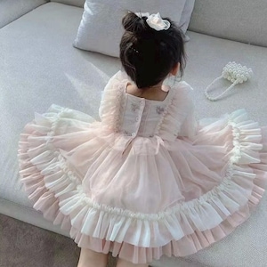 May include: A pale pink and white ruffled dress with a square neckline and long sleeves. The dress features a layered skirt and decorative buttons at the back. A white flower hair accessory is visible.