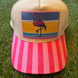 May include: A beige baseball cap featuring a rectangular patch with a pink flamingo graphic. The patch has horizontal stripes in blue, yellow, and teal. The brim of the cap displays vertical stripes in alternating shades of bright and light pink.