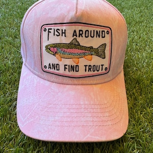 Può includere: Un cappellino da baseball rosa chiaro con una texture sbiadita. Al centro è presente una toppa rettangolare con una trota iridea ricamata e il testo nero "FISH AROUND AND FIND TROUT" con piccoli cerchi agli angoli.