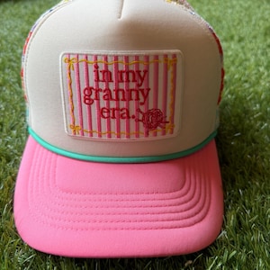 May include: A trucker hat with a cream-colored front panel, a pink brim, and a teal cord trim. The front features a rectangular patch with pink vertical stripes and the embroidered red text "in my granny era." with a small yarn ball graphic.