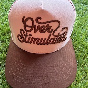 May include: A light pink baseball cap with a dark brown brim and a small dark brown button on top. The front panel features the text Over Stimulated embroidered in dark brown thread with a cursive, flowing script style font and stitching.