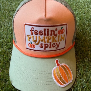 Könnte beinhalten: Eine pfirsich- und salbeigrüne Trucker-Cap mit einem rechteckigen Aufnäher, der den gestickten Text 'feelin' PUMPKIN spicy' in Orange und Braun zeigt, ergänzt durch zwei kleine Kürbis-Symbole und einen großen gestickten Kürbis-Aufnäher auf dem Schirm.