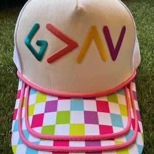 May include: A cream-colored trucker hat with a white mesh back and a pink button on top. The front features the embroidered letters "G>AV" in teal, red, yellow, and purple. The brim has a colorful checkered pattern with pink trim.