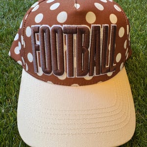 Puede incluir: Gorra de béisbol marrón y beige con la palabra "FOOTBALL" bordada en marrón. La corona de la gorra presenta un estampado de lunares y la visera es de color beige liso. La gorra está sobre hierba verde.