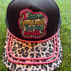 May include: A black trucker hat with a mesh back and a leopard print brim accented by pink piping. The front panel features a colorful embroidered patch with the text "I have a good heart, But this mouth..." in stylized lettering with pink and teal details.