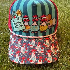 May include: A trucker hat featuring a teal and dark teal striped front panel with embroidered patches of three chickens, a tote bag labeled FRESH, and eggs. The brim has a repeating pattern of white chickens and red stars on a dark blue background.