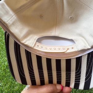 Pu&ograve; includere: Un cappellino da baseball beige con visiera a righe bianche e nere. Il cappellino ha un cinturino regolabile beige e una visiera curva. Il lato inferiore della visiera presenta strisce verticali bianche e nere.