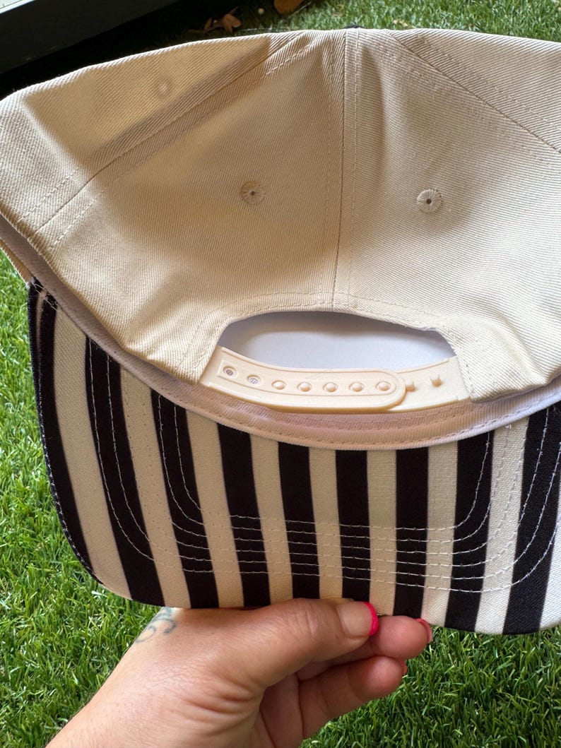 Pu&ograve; includere: Un cappellino da baseball beige con visiera a righe bianche e nere. Il cappellino ha un cinturino regolabile beige e una visiera curva. Il lato inferiore della visiera presenta strisce verticali bianche e nere.