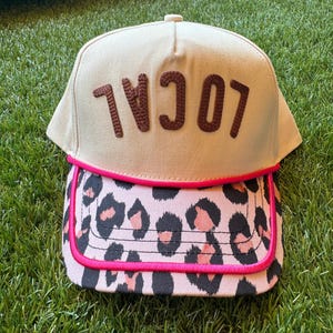 May include: A beige baseball cap with the word "LOCAL" embroidered in brown. The brim has a pink leopard print design with a hot pink trim. The cap is on green grass.