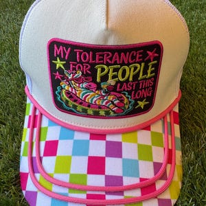 May include: A white trucker hat with a mesh back and a pink button on top. The front features a rectangular patch with a zebra wearing sunglasses in a pool float, and the text: MY TOLERANCE FOR PEOPLE LAST THIS LONG. The brim has a colorful checkerboard pattern.