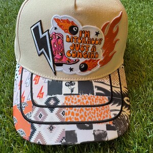 May include: A beige baseball cap featuring a large embroidered patch with a pink cowboy boot, two flaming billiard balls, and the text I'M LITERALLY JUST A COWGIRL. The brim displays a multi-patterned print including leopard spots, checkers, and lightning bolts.