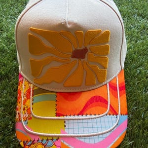 Op de afbeelding: Een beige honkbalpet met een geel geborduurd abstract bloemmotief op het voorpaneel. De klep heeft een veelkleurig patchworkpatroon met oranje, roze, blauwe en gele bloemen- en rastermotieven, geaccentueerd door witte stiksels.
