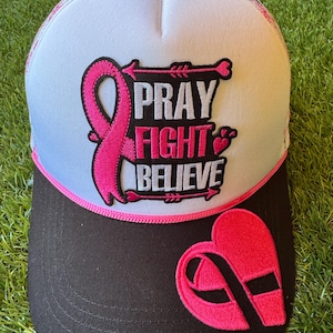 Peut inclure: Une casquette trucker blanche et noire avec un ruban rose et le texte PRAY FIGHT BELIEVE brodé en blanc et rose sur le panneau avant. La visière est noire avec un écusson en forme de cœur rose et ruban noir, et un cordon rose sépare les panneaux.