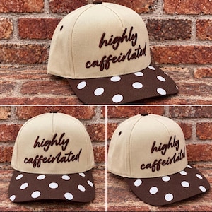 Pode incluir: Um boné de beisebol bege com o texto bordado em castanho escuro "highly caffeinated" no painel frontal. O boné tem uma aba castanha escura decorada com um padrão repetitivo de bolinhas brancas e um pequeno botão castanho escuro no topo.
