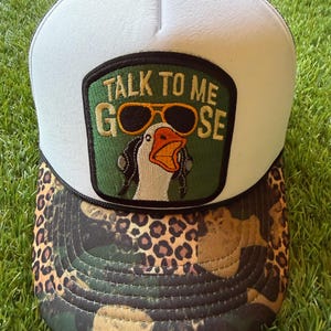 May include: A white trucker hat with a leopard and camouflage print brim. The front panel features a rectangular patch with a green background, an embroidered goose wearing yellow sunglasses and headphones, and the text TALK TO ME GOOSE.
