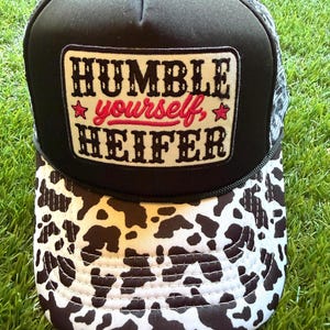 May include: A black trucker hat featuring a white rectangular patch with the black text HUMBLE and HEIFER, and the red script text yourself, flanked by two red stars. The brim has a black and white cow print pattern, and the back is mesh.