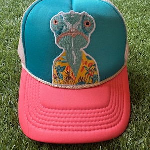 May include: A teal and coral trucker hat featuring an embroidered patch of a chameleon wearing a yellow floral shirt. The cap has a mesh back, a solid front panel, and a braided cord trim separating the crown from the curved coral brim.