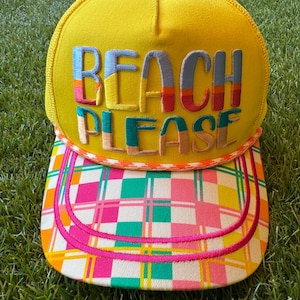 May include: A bright yellow trucker cap featuring the embroidered text BEACH PLEASE in multi-coloured block letters. The cap has a structured front panel, a mesh back, and a flat brim with a vibrant pink, yellow, green, and white plaid pattern.