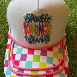 Può includere: Un cappellino trucker bianco con un bottone rosa sulla parte superiore e un cordoncino rosa lungo la visiera. La parte anteriore presenta una toppa ricamata con il testo Sprinkle KINDNESS everywhere in lettere multicolori. La visiera è a scacchi.