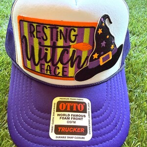 May include: A purple and white trucker hat featuring a rectangular patch with the text "RESTING WITCH FACE" in black lettering, an orange broomstick graphic, and a black witch hat with yellow stars. A sticker on the brim reads "OTTO".