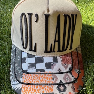 May include: A beige baseball cap featuring the black embroidered text OL' LADY on the front crown. The brim displays a multi-patterned patchwork design including black and white checkers, orange leopard print, and geometric shapes in muted tones.