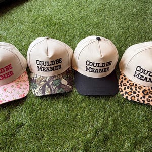 Può includere: Quattro cappellini da baseball con corona beige e il testo ricamato COULD BE MEANER. Il primo ha una visiera a fiori rosa, il secondo una visiera mimetica, il terzo una visiera nera tinta unita e il quarto una visiera leopardata.