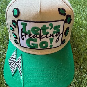 Puede incluir: Una gorra de béisbol beige con visera verde. El panel frontal presenta un parche rectangular blanco con el texto verde y negro "Let's freaking Go!" rodeado de manchas de leopardo verdes y negras. Un parche de rayo con estampado animal en blanco y negro está en la visera.