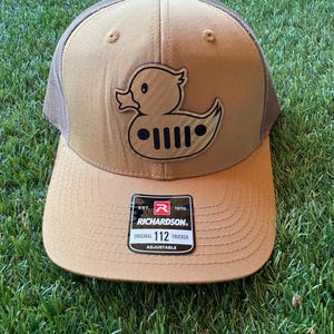 Peut inclure: Casquette de camionneur beige et marron avec un logo de canard représentant une calandre Jeep. La casquette a une visière incurvée et un dos en filet. L'étiquette de la marque Richardson est visible.